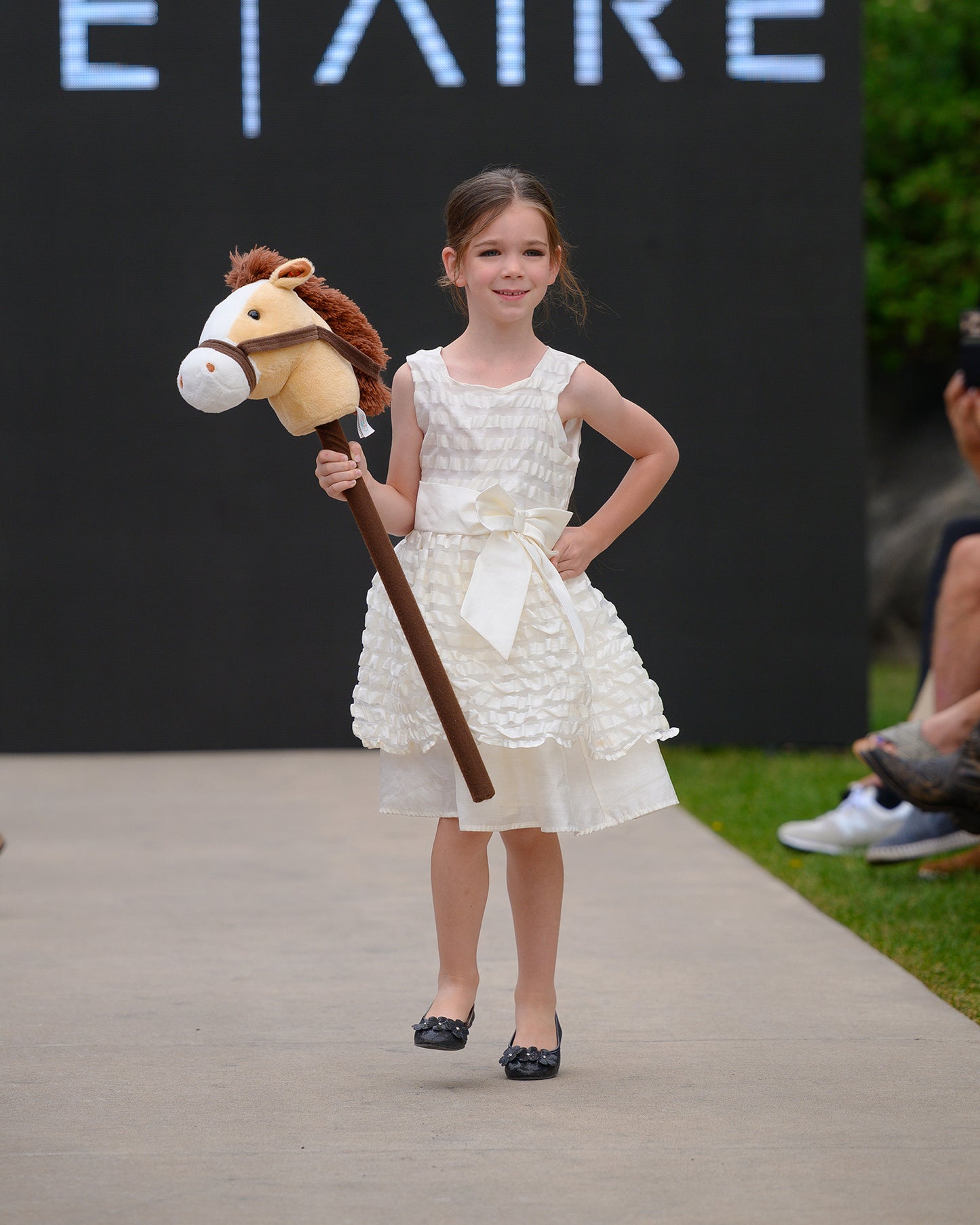 Young girl in a white dress holding a toy horse on a runway with branding in the background.