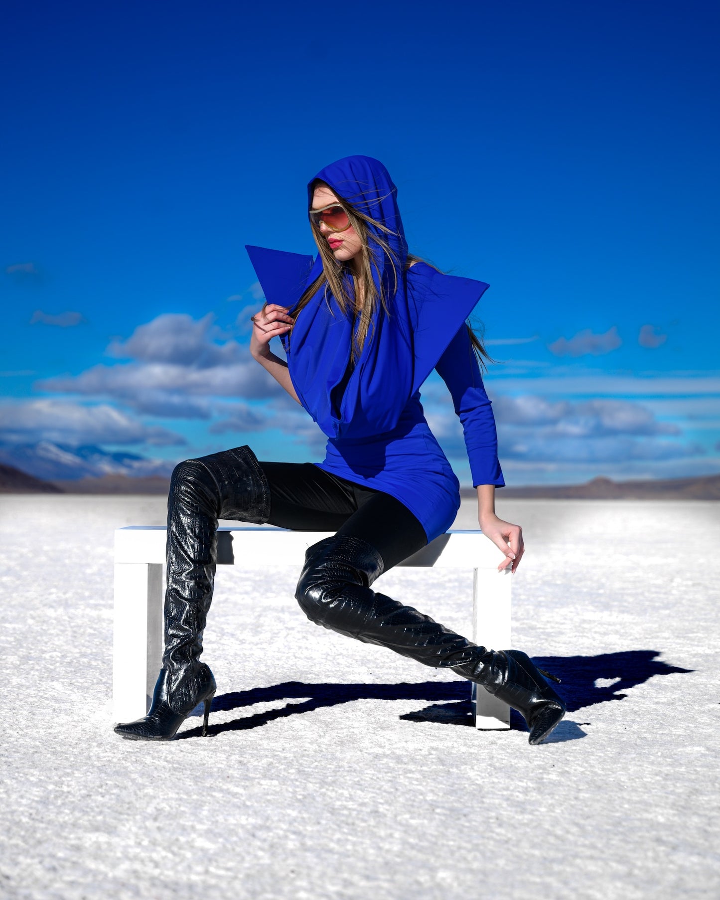 Model wearing a blue outfit sitting on a white block in a desert landscape with blue sky.