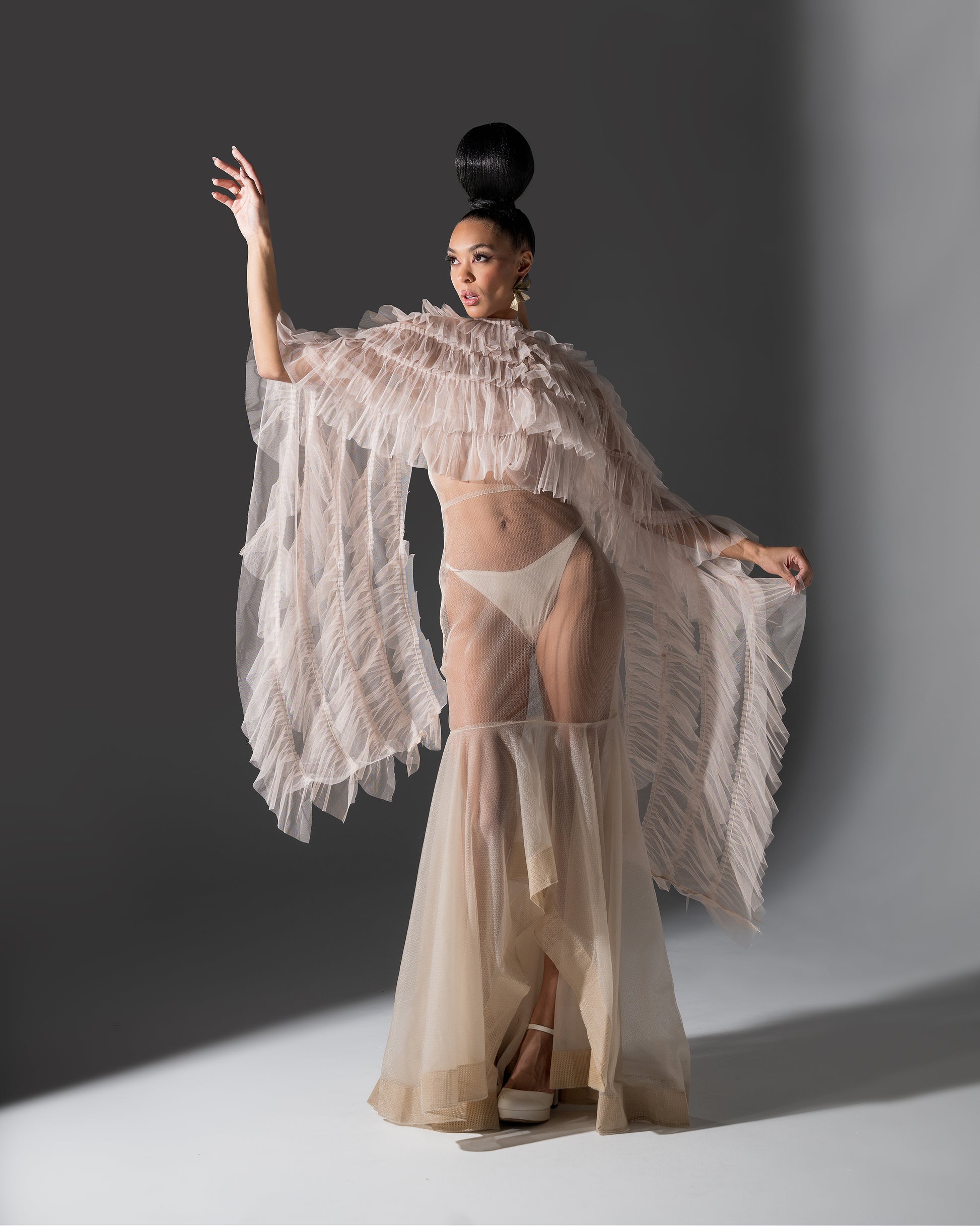 Model wearing a sheer beige outfit with a white ruffled cover-up against a gray background